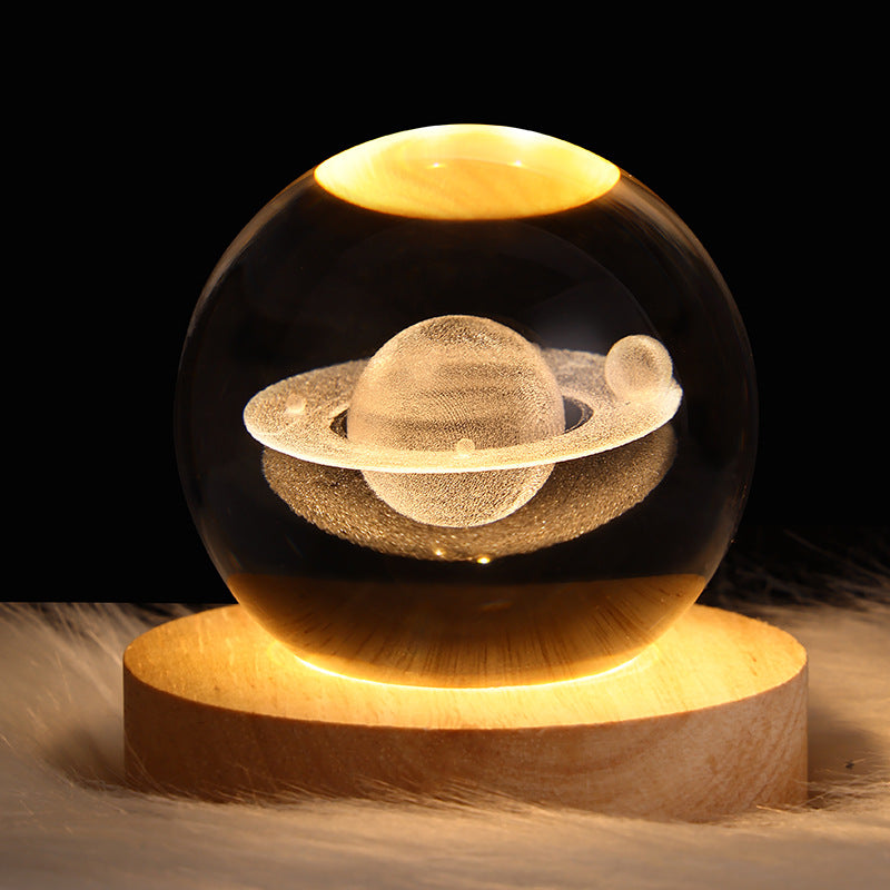 A clear glass crystal ball lamp featuring an etched Saturn design, resting on a round wooden base that glows with warm light against a black background.