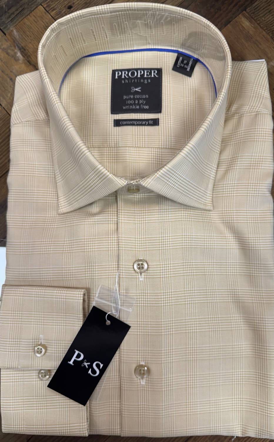 A folded beige dress shirt with a subtle windowpane check pattern, featuring a collar label for 'PROPER Suits' and a black 'P+S' hangtag on the cuff, displayed on a dark wood background.
