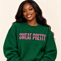 A woman with long dark hair wearing a green crewneck sweatshirt with the text 'SWEAT PRETTY SWEAI PRETTY' printed in pink across the chest, set against a plain beige background.