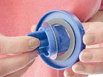 A close-up of a person's hands holding a blue plastic baby bottle nipple or breast shield attachment against a pink background.