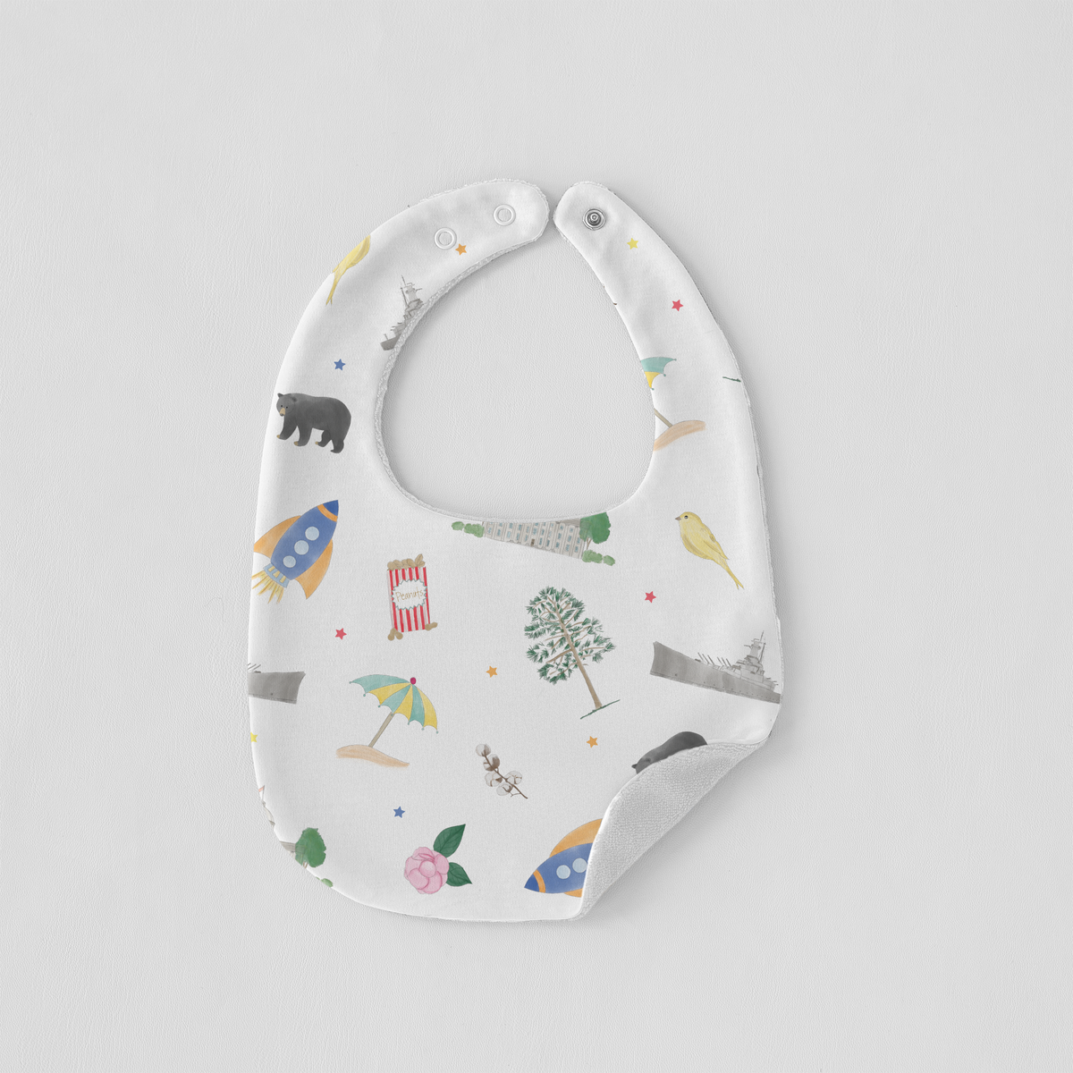 A white baby bib with a colorful whimsical print of rockets, a circus tent, a tree, a bird, and stars, shown on a light gray background.