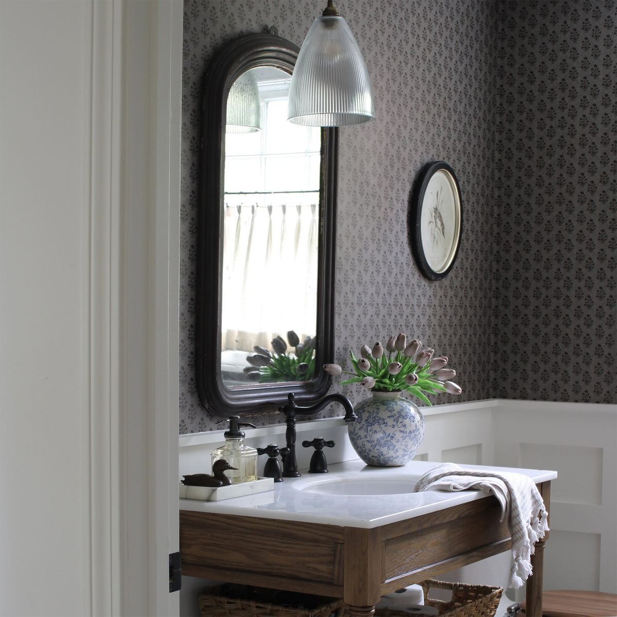 A styled bathroom interior featuring dark gray patterned wallpaper, a dark wood vanity with a white sink, a black faucet, and a blue-and-white ceramic vase holding a succulent.