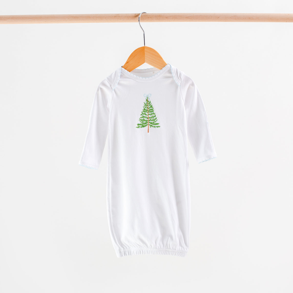 A white long-sleeve baby bodysuit or sleep sack hanging on a wooden hanger from a wooden rod, featuring a small green Christmas tree graphic on the chest.