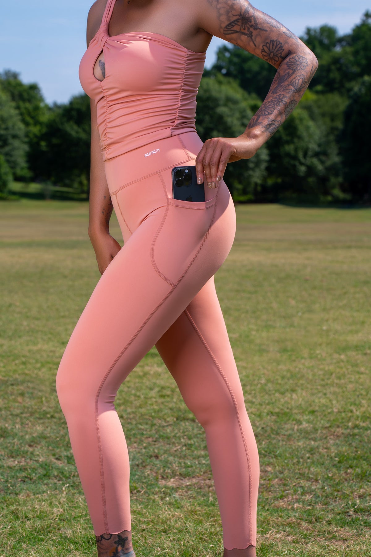 Women putting phone in her pocket wearing peach one shoulder Key hole athletic shirt and matching leggings with outdoor field background with trees.  