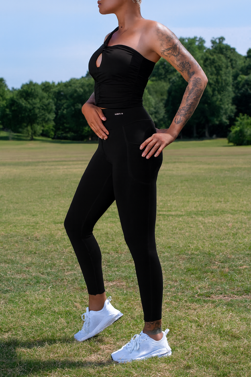 A woman modeling a black strapless athletic top and black leggings with white sneakers, standing in a grassy park with trees and blue sky in the background.