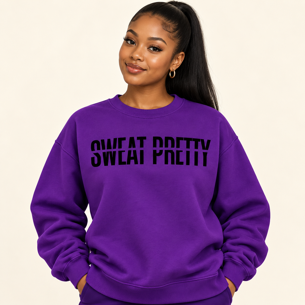 A woman with a high ponytail wearing a purple crewneck sweatshirt with the text 'SWEAT PRETTY' printed in black across the chest, posed against a solid light beige background.