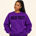 A woman with a high ponytail wearing a purple crewneck sweatshirt with the text 'SWEAT PRETTY' printed in black across the chest, posed against a solid light beige background.