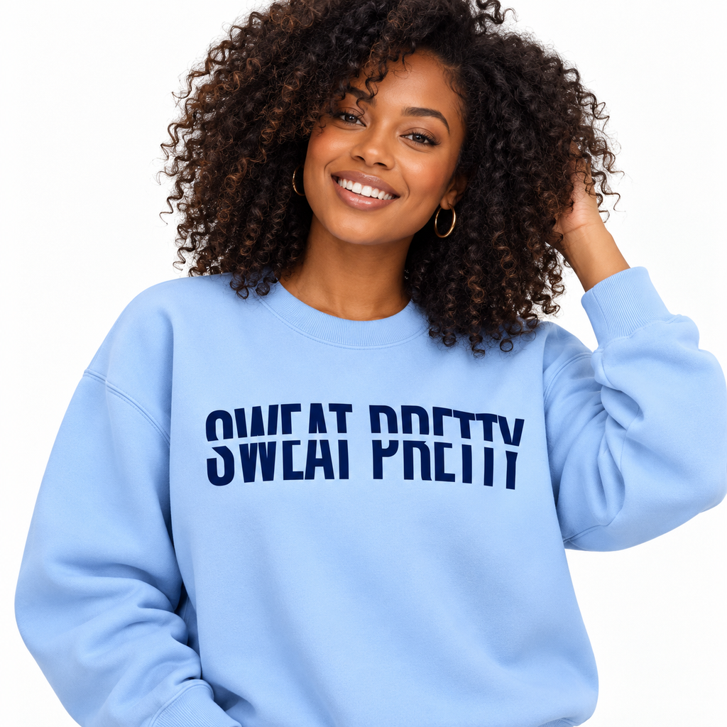 A smiling woman with curly dark hair wears a light blue crewneck sweatshirt with the text 'SWEAT PRETTY' printed in dark blue across the chest, set against a plain white background.
