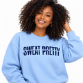 A smiling woman with curly dark hair wears a light blue crewneck sweatshirt with the text 'SWEAT PRETTY' printed in dark blue across the chest, set against a plain white background.