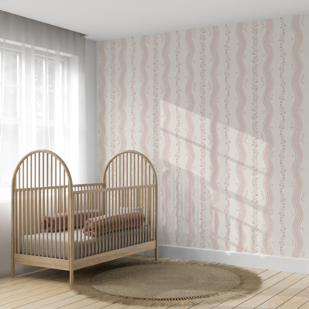 A nursery room scene featuring a light wood crib with arched headboard and footboard, styled with a brown blanket and mattress. The wall behind the crib is covered in a vertical patterned wallpaper with wavy stripes in dusty pink and beige.