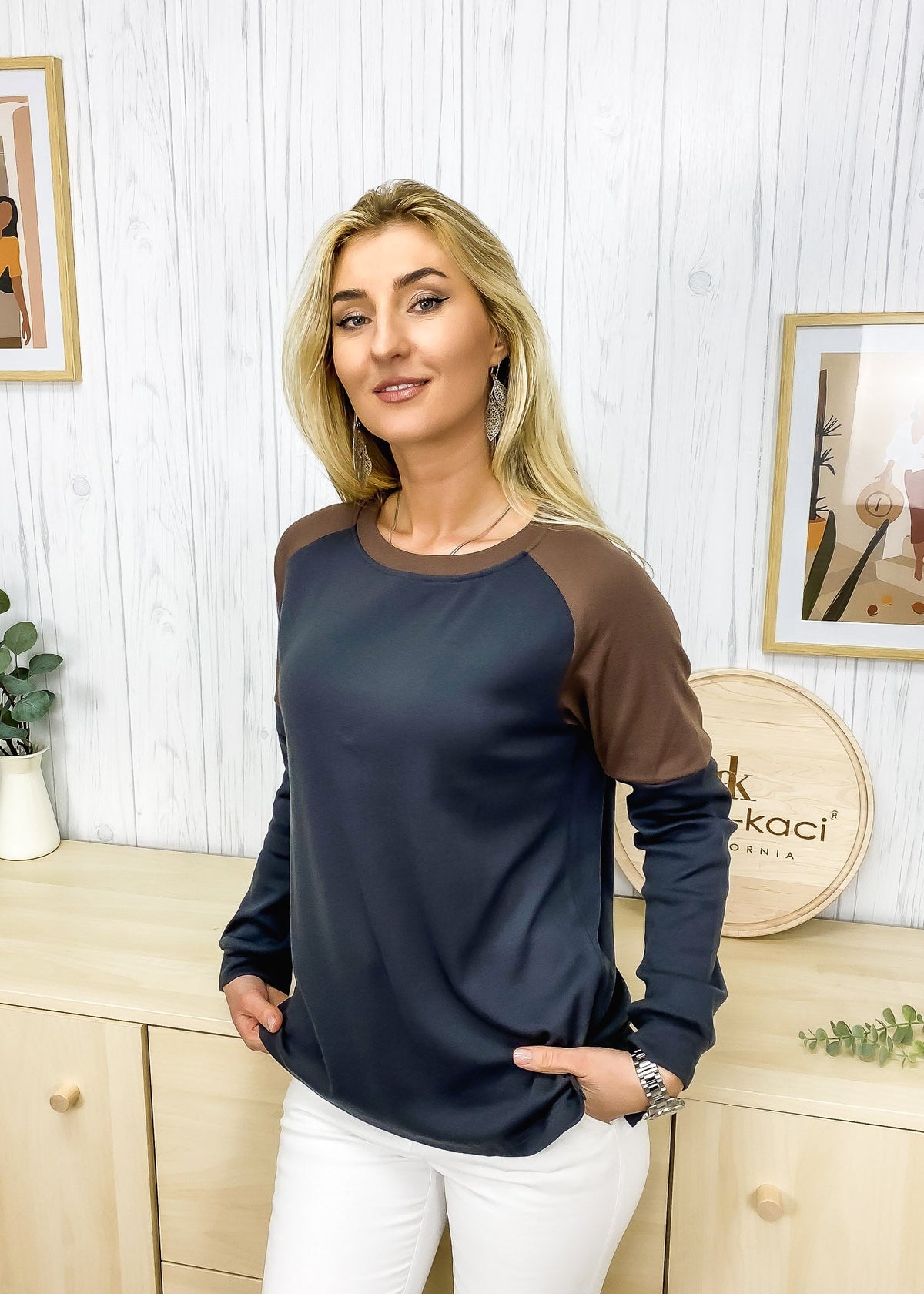 Woman wearing a navy and brown long-sleeve top in a room with wooden furniture and decorative items.