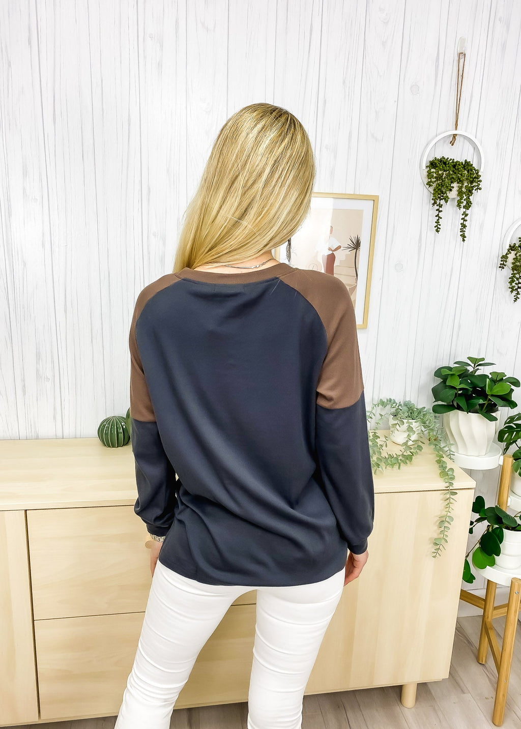 Woman wearing a navy and brown long-sleeve top in a room with plants and furniture.