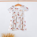 A short-sleeve white dress with a cartoon dog print, hanging on a wooden hanger against a white wall with dried grass at the bottom.