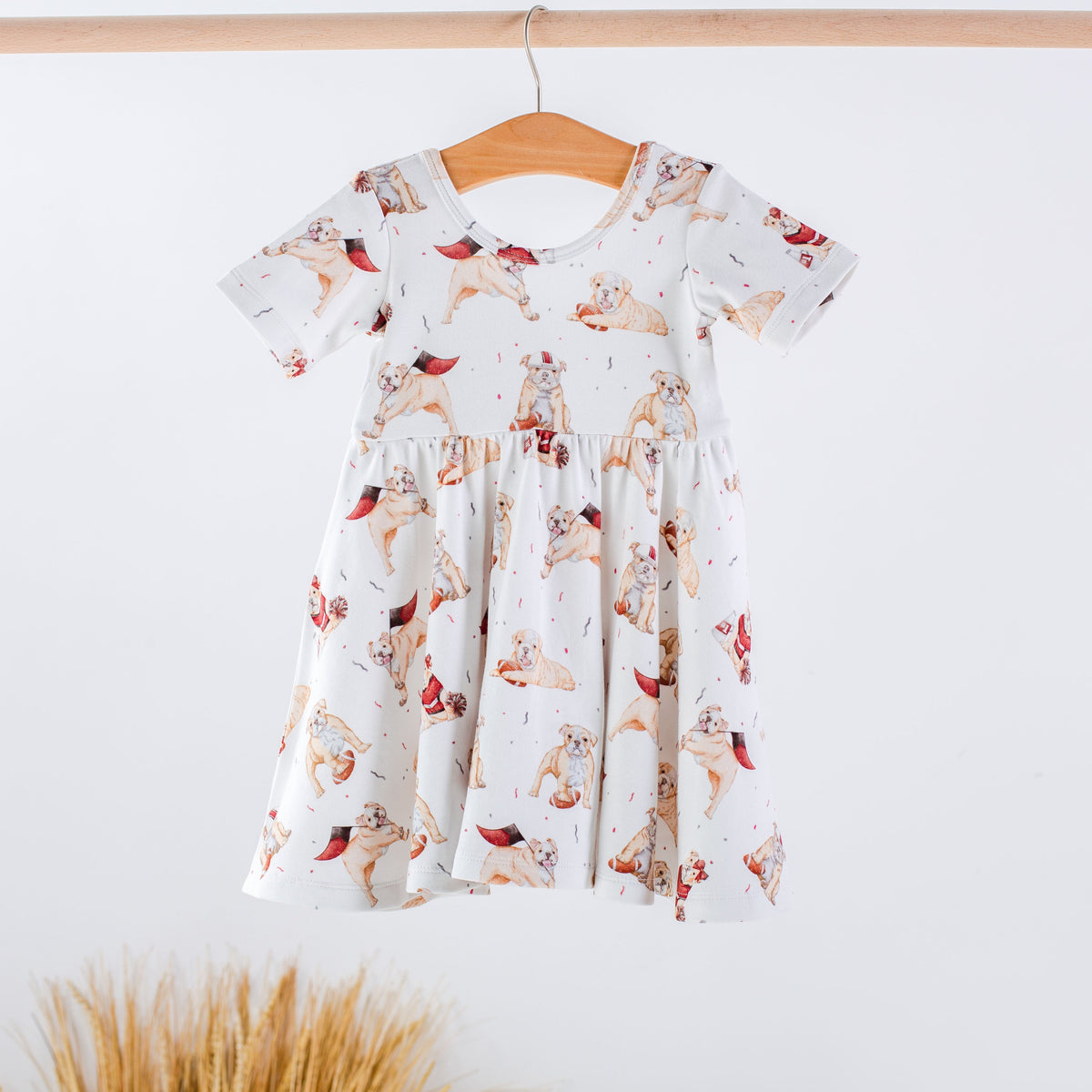 A short-sleeve white dress with a cartoon dog print, hanging on a wooden hanger against a white wall with dried grass at the bottom.