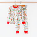 A children's two-piece pajama set hanging on wooden hangers against a plain white wall. The set includes a long-sleeve top and matching pants in white with a colorful Christmas-themed print featuring Santa Claus, reindeer, candy canes, and holiday motifs, finished with red ribbed cuffs and collar.