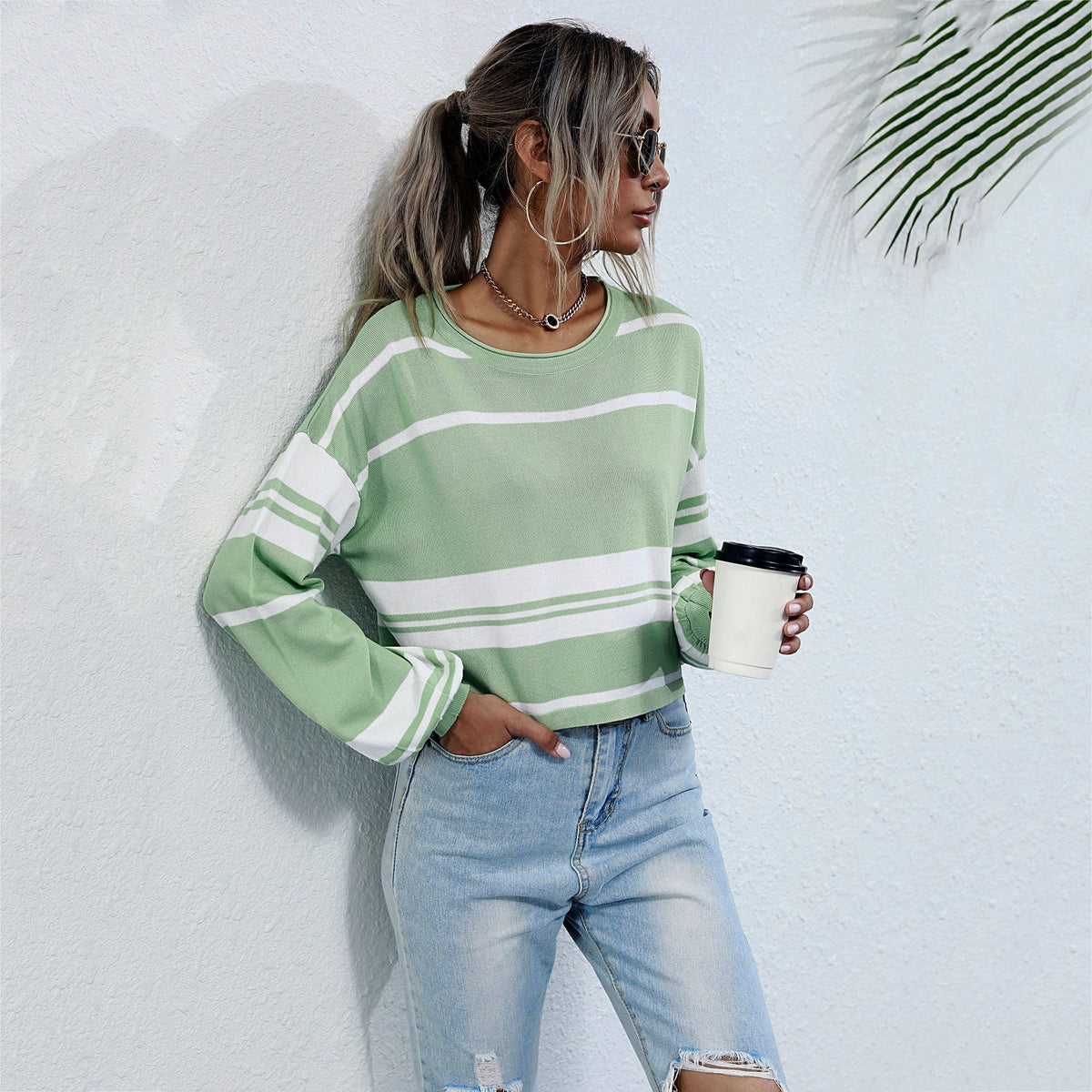 Woman wearing a green and white striped sweater and blue jeans, holding a coffee cup against a light background.