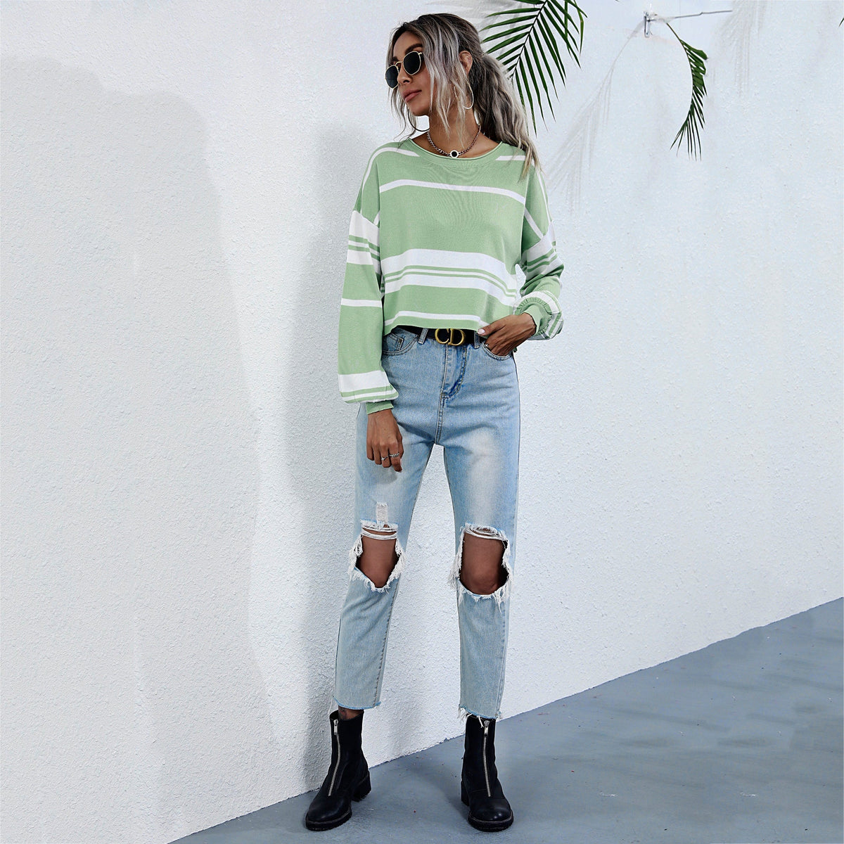 Woman wearing a green and white striped sweater with ripped jeans against a white wall.