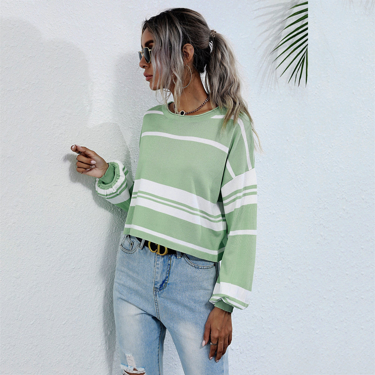 Woman wearing a green and white striped sweater and blue jeans against a white wall.