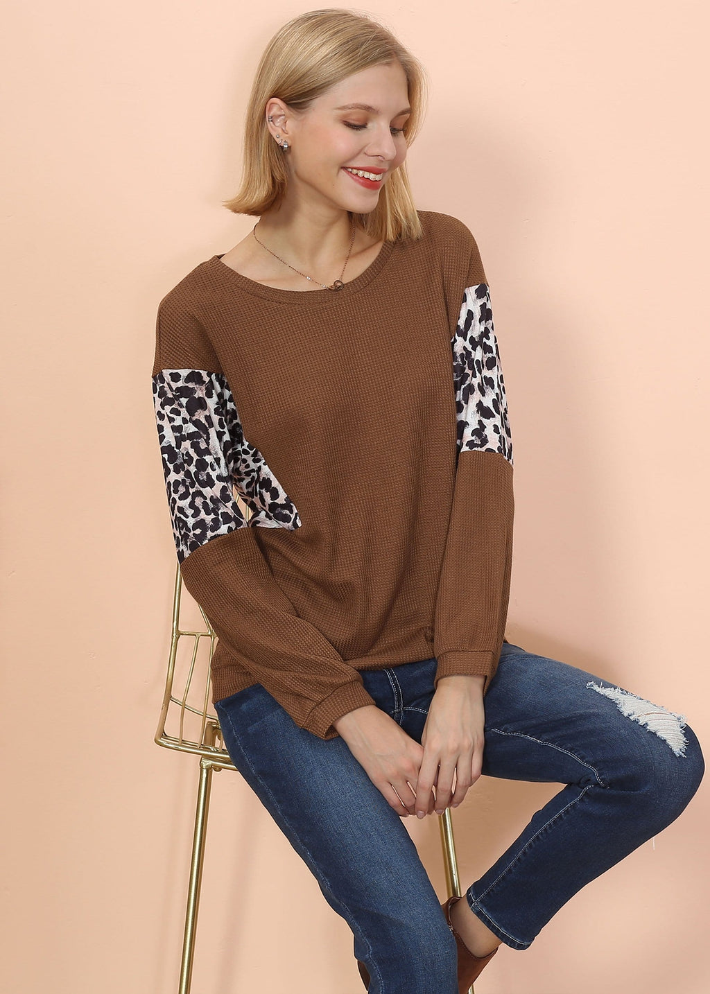 Woman wearing a brown sweater with leopard print sleeves sitting on a chair against a beige background