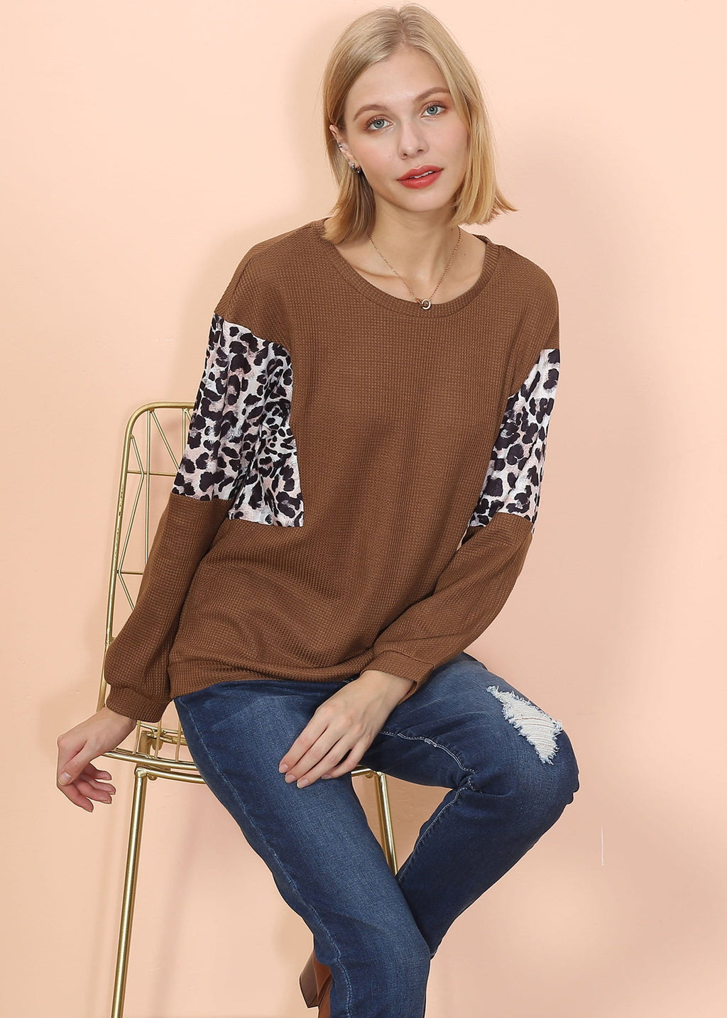 Woman wearing a brown sweater with leopard print sleeves sitting on a chair against a beige background