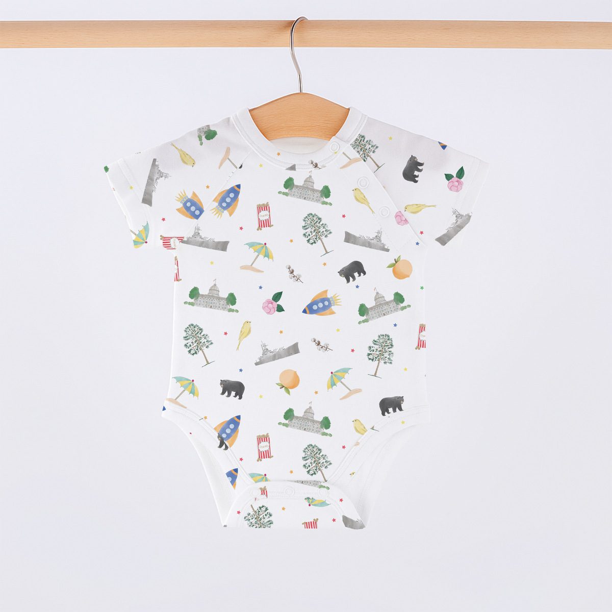 A white short-sleeve baby bodysuit with an all-over whimsical print of cartoon animals, trees, stars, and small objects, hanging on a wooden hanger from a wooden rod against a light gray background.