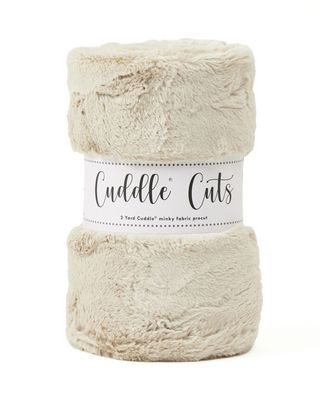 A folded plush faux fur fabric in a light oatmeal beige color, wrapped in a white paper band that reads 'Cuddle Cuts'.