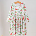 A long-sleeve children's dress hanging on a wooden hanger from a clothing rack. The dress has a white base with a colorful Christmas-themed print featuring red Santa Cabs, green pine trees, and red Christmas trees.