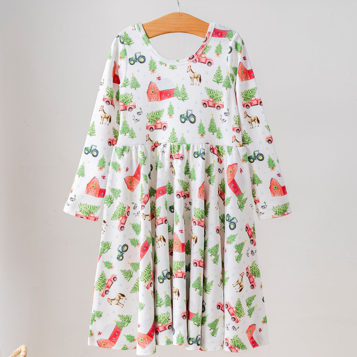 A long-sleeve children's dress hanging on a wooden hanger from a clothing rack. The dress has a white base with a colorful Christmas-themed print featuring red Santa Cabs, green pine trees, and red Christmas trees.