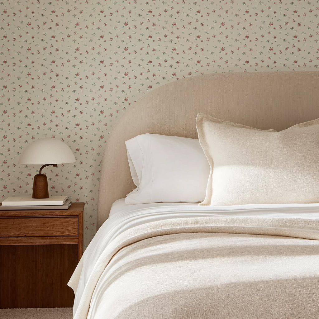 A styled bedroom scene featuring a bed with cream-colored bedding, a beige upholstered headboard, and a wall covered in delicate floral-patterned wallpaper. A wooden nightstand with a white table lamp sits on the left side.