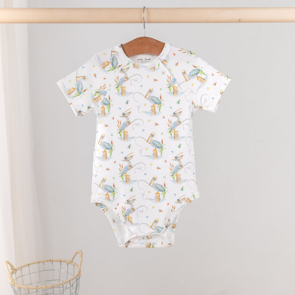 A baby short-sleeve bodysuit with an all-over print of blue birds, hanging on a wooden hanger from a wooden clothes rack. The bodysuit is white with a pattern of light blue birds, yellow acorns, and small gray tree trunks.