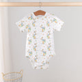 A baby short-sleeve bodysuit with an all-over print of blue birds, hanging on a wooden hanger from a wooden clothes rack. The bodysuit is white with a pattern of light blue birds, yellow acorns, and small gray tree trunks.