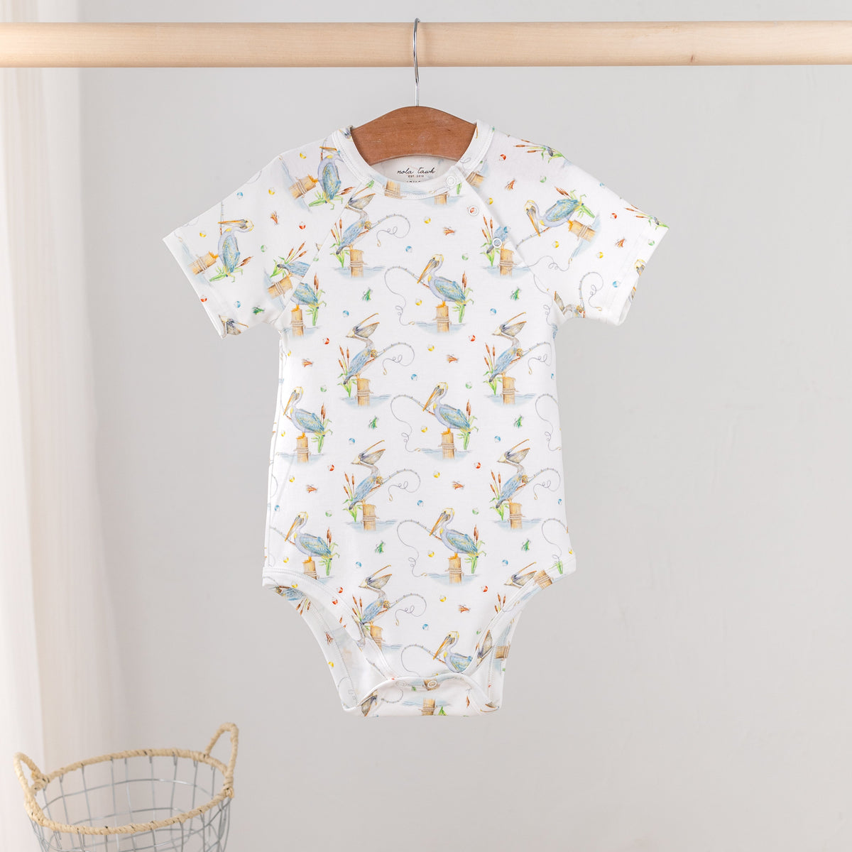 A baby short-sleeve bodysuit with an all-over print of blue birds, hanging on a wooden hanger from a wooden clothes rack. The bodysuit is white with a pattern of light blue birds, yellow acorns, and small gray tree trunks.