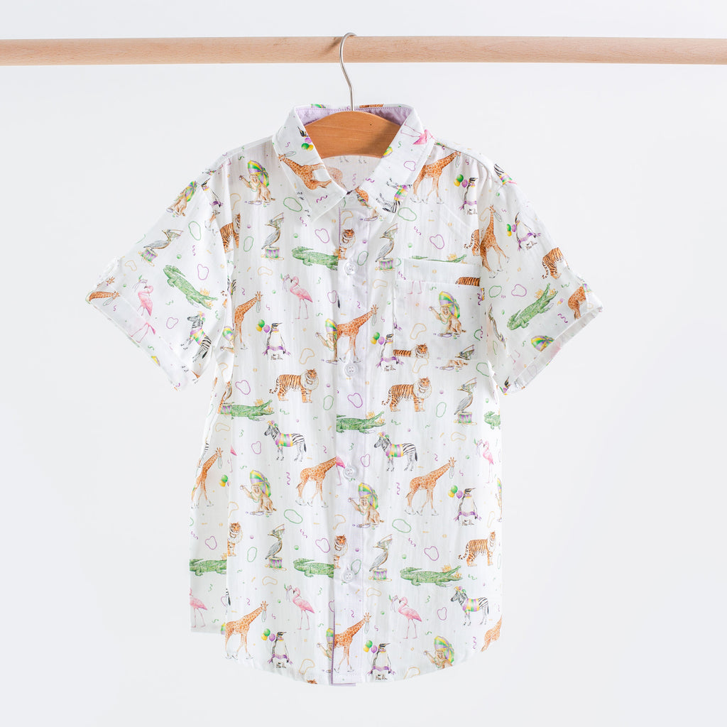 A short-sleeve button-up shirt hanging on a wooden hanger against a light gray background. The shirt has a white base with a colorful cartoon print of giraffes, crocodiles, cactus plants, and small hearts.