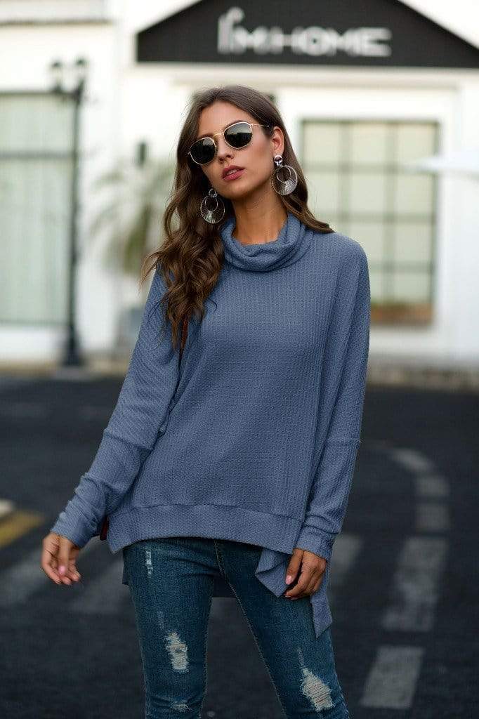Woman wearing a blue sweater and jeans with sunglasses, standing on a street.