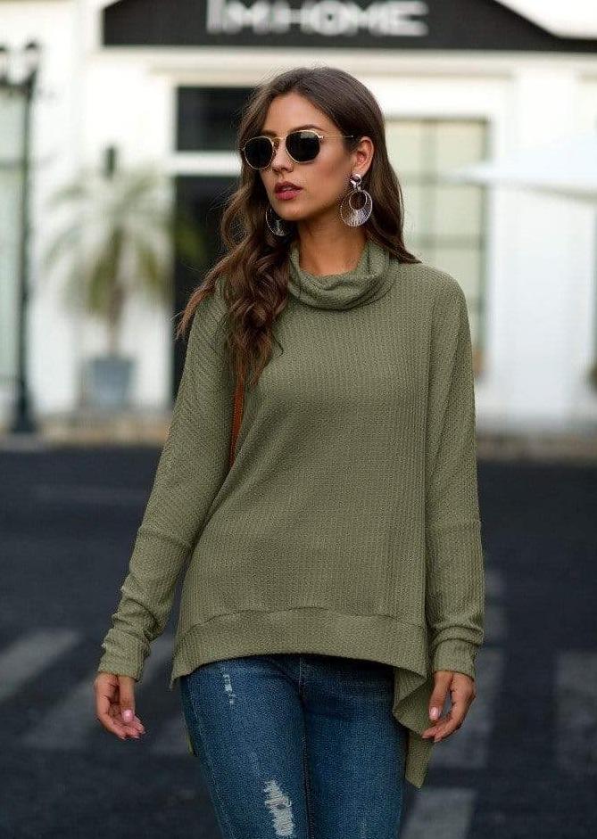 A woman wearing an olive green waffle-knit cowl-neck sweater and blue jeans, standing outdoors in front of a blurred building background.